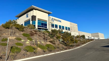 Image of a white industrial building's facade from the ground, showing the green landscaping and the DrinkPAK logo on the front of the building